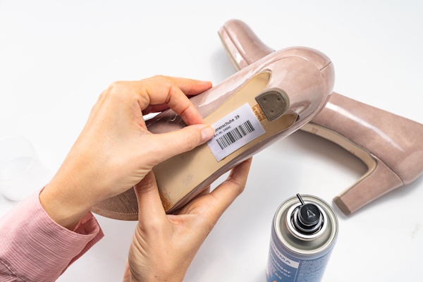 Une main tient une chaussure avec une étiquette de prix, tandis qu'une bombe de spray se trouve à côté. La chaussure est un escarpin beige.