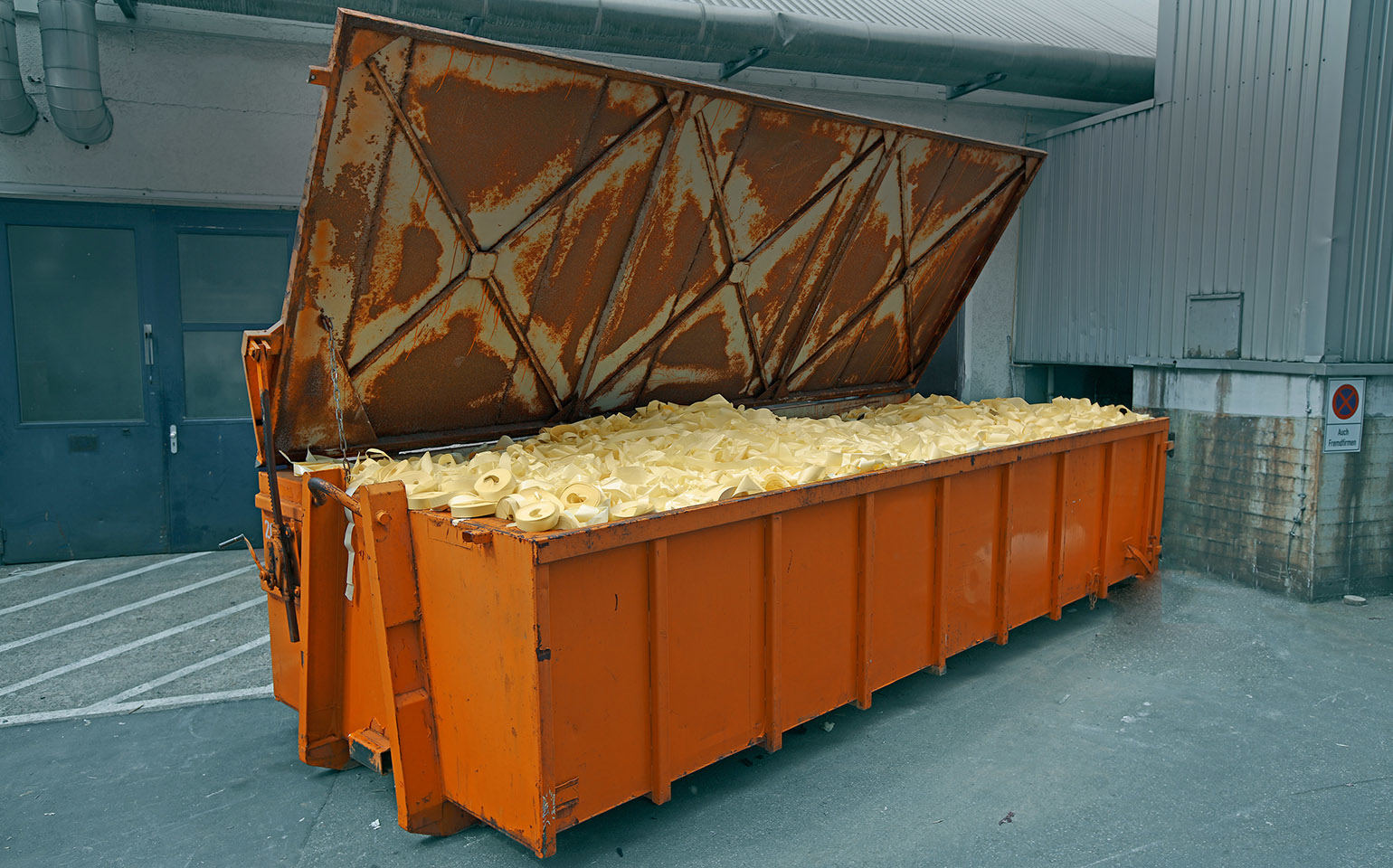 Un grand conteneur orange rempli de déchets jaunes est adossé à un mur, avec un couvercle ouvert.