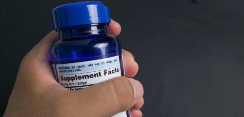 Une main tient une bouteille bleue de compléments alimentaires, sur l'étiquette figurent les informations sur les suppléments.