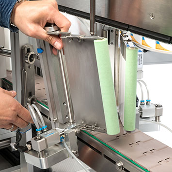 Une main ajuste une plaque métallique sur une machine, tandis que deux rouleaux verts sont visibles sur le côté droit.