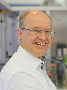 Un homme avec des lunettes et un visage souriant se tient dans un environnement de laboratoire, entouré d'appareils techniques et d'équipements.