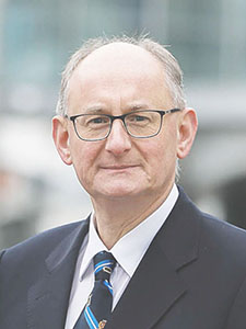 Un homme portant des lunettes et un costume se tient à l'extérieur. Il a des cheveux gris et porte une cravate rayée.