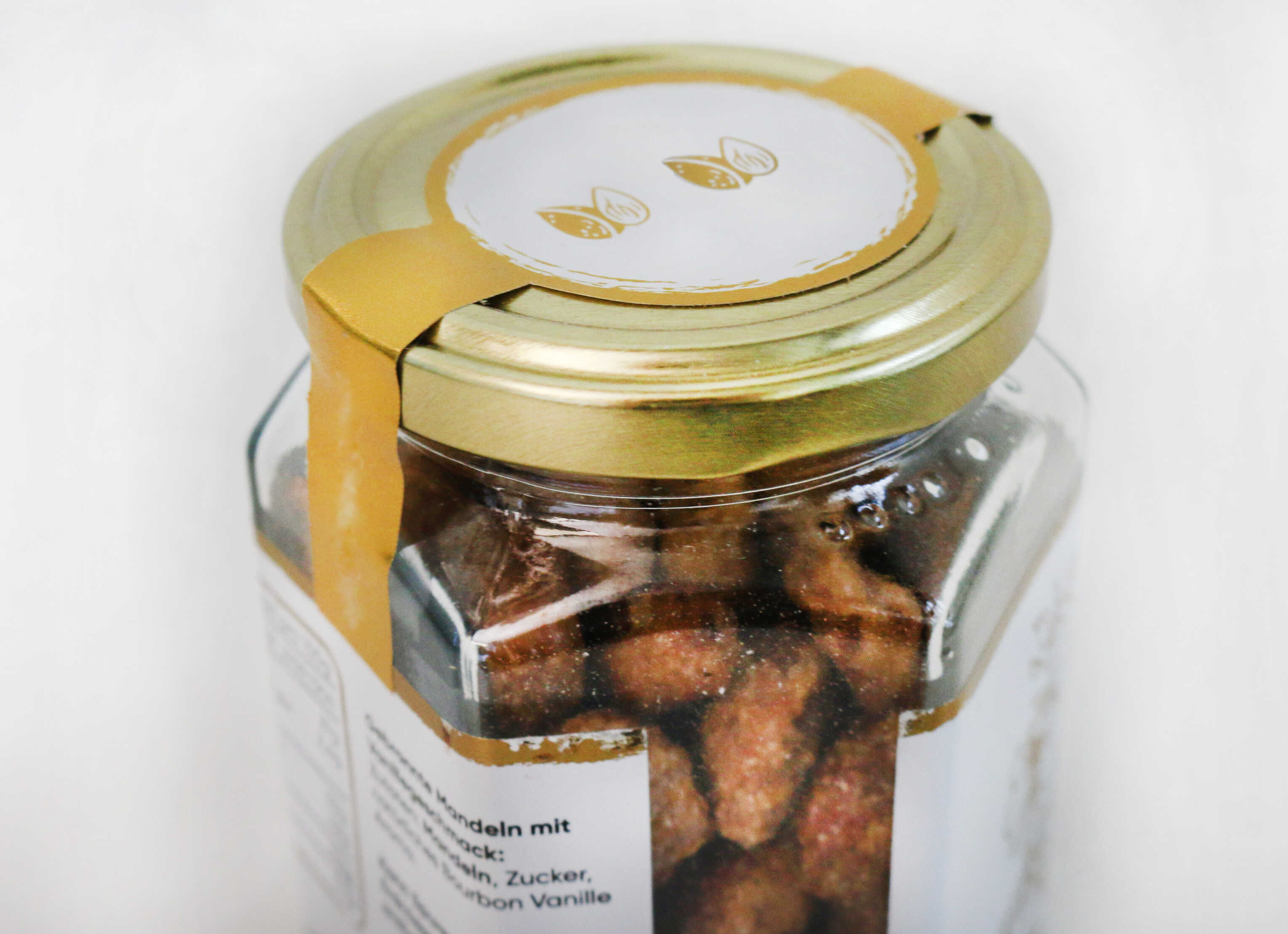 Un verre avec un couvercle doré, rempli d'amandes grillées. Sur le couvercle, il y a une étiquette au design minimaliste.