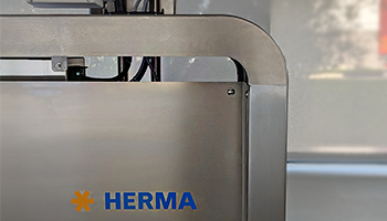 Une partie d'une machine avec le logo 'HERMA' en lettres bleues sur une surface métallique.