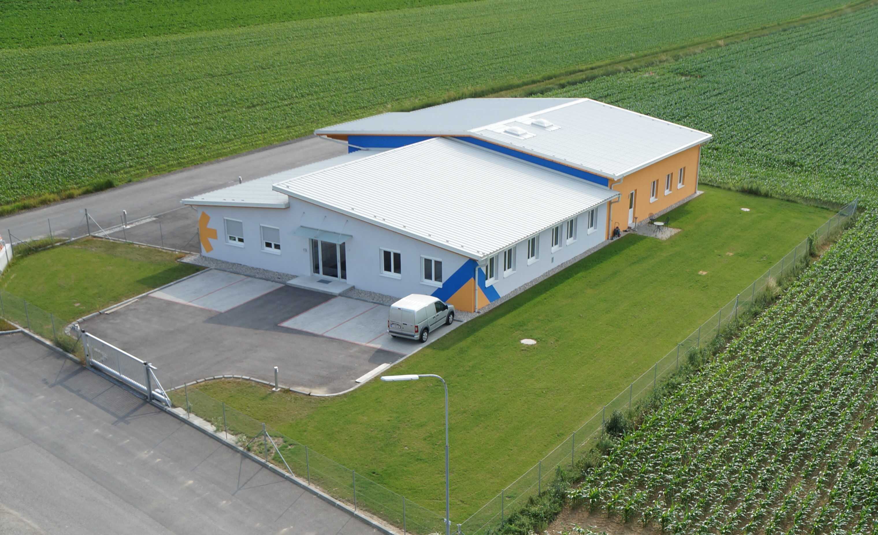 Un bâtiment moderne avec un toit plat, une façade lumineuse et des accents colorés, entouré de champs agricoles.