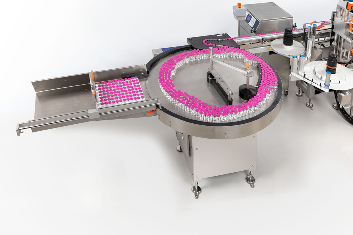 Installation de remplissage automatisée avec un convoyeur circulaire, rempli de petites bouteilles. Des étiquettes roses sont visibles.