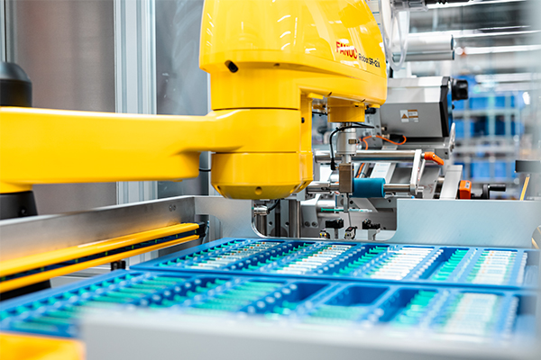 Un bras de robot industriel jaune travaille sur un tapis roulant avec des conteneurs bleus remplis de divers objets.