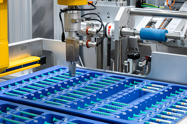 Machine automatisée traitant des seringues dans des conteneurs bleus, avec un bras robotisé qui saisit une seringue.