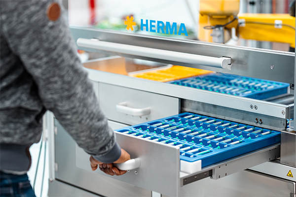 Une personne tire un tiroir avec des compartiments bleus d'une étagère marquée du logo "HERMA".