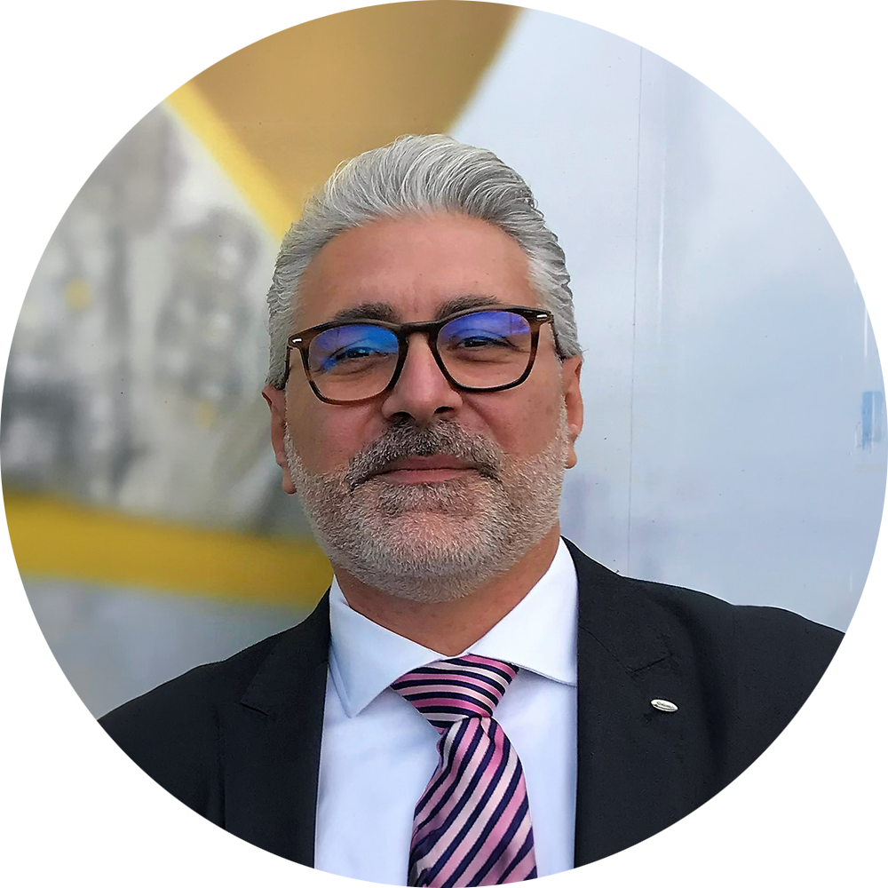 Homme aux cheveux gris et portant des lunettes, vêtu d'un costume avec une cravate rayée. Arrière-plan flou et lumineux.