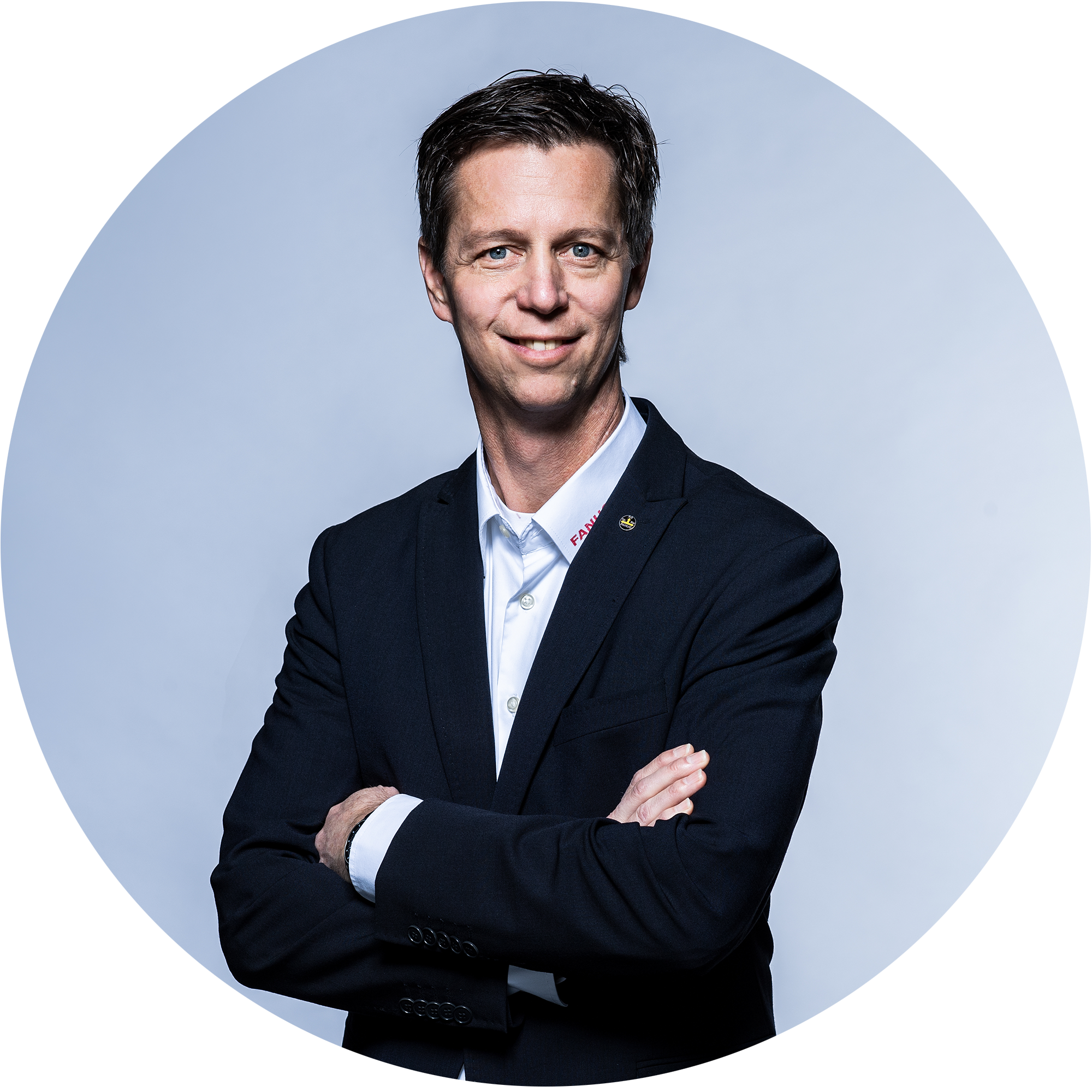 Un homme en costume noir, les bras croisés, sourit à la caméra. L'arrière-plan est clair et neutre.