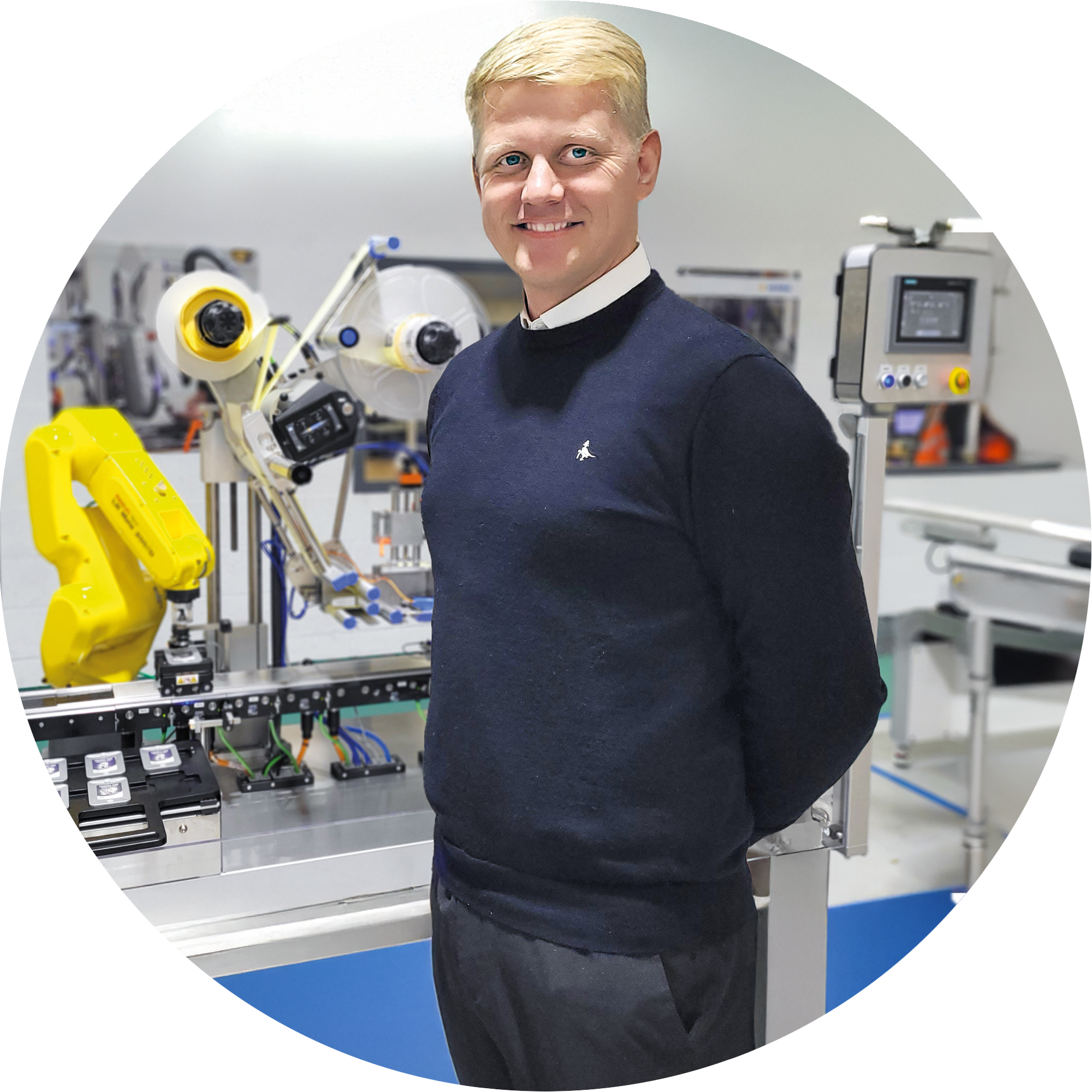 Un homme en pull noir se tient à côté d'une ligne de production automatisée avec des robots et des équipements techniques.