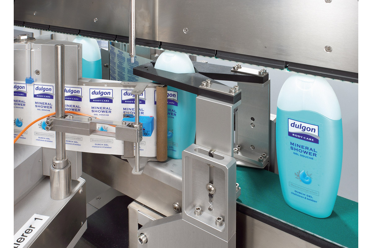 Une bouteille de Dulgon Mineral Shower est posée sur un tapis roulant, entourée d'emballages. Des composants de machines sont visibles.