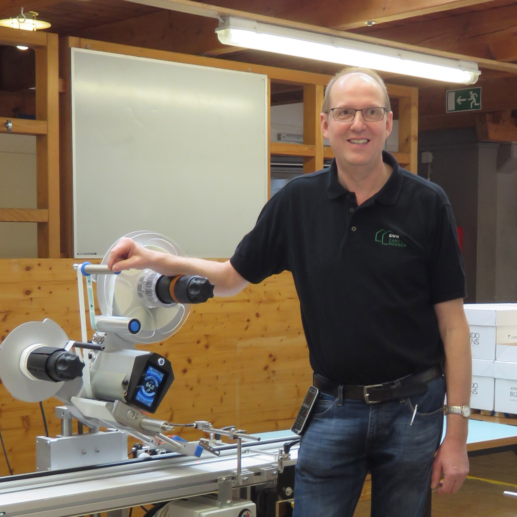 Un homme se tient à côté d'une machine dans un atelier, sourit et porte un t-shirt noir avec un logo.
