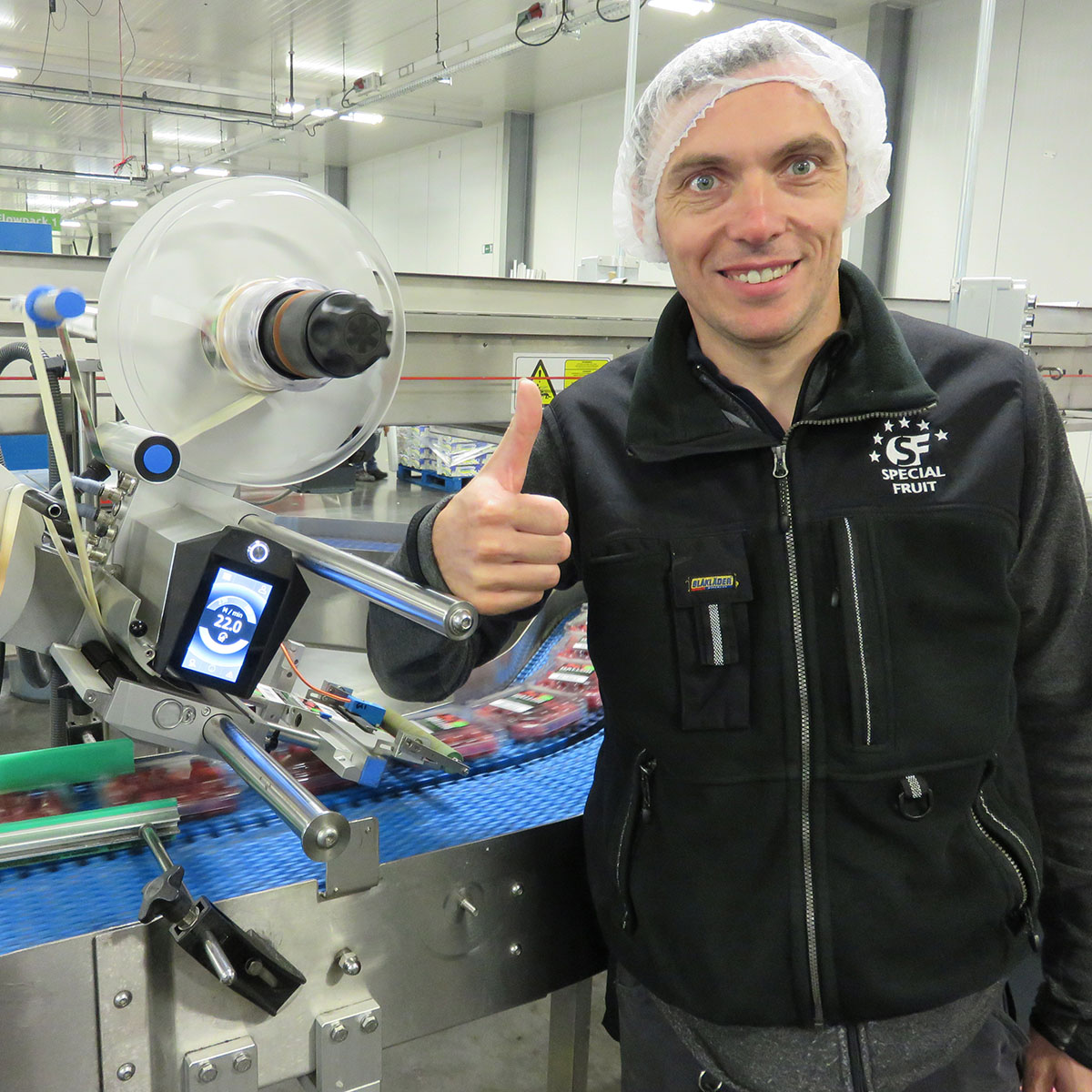 Homme avec un pouce levé dans une usine, à côté d'une machine à étiqueter des produits sur un tapis roulant.