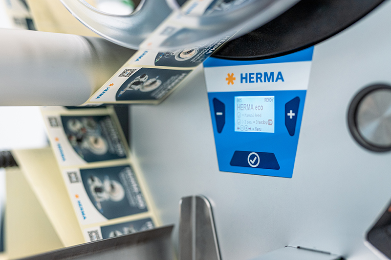 Un imprimante d'étiquettes avec un écran affichant la marque HERMA. Les étiquettes sont délivrées sur un rouleau.