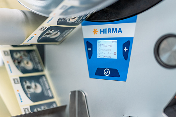 Un imprimante d'étiquettes avec un panneau de commande numérique affichant la marque "HERMA". Les rouleaux d'étiquettes sont visibles.