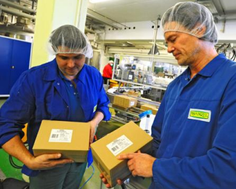 Deux ouvriers en salopettes bleues et filets à cheveux tiennent des cartons dans un environnement de production et vérifient leur contenu.