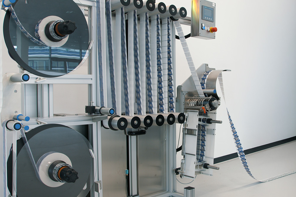 Une machine industrielle avec deux grandes bobines qui fait passer un ruban à travers plusieurs rouleaux, équipée d'un panneau de commande.