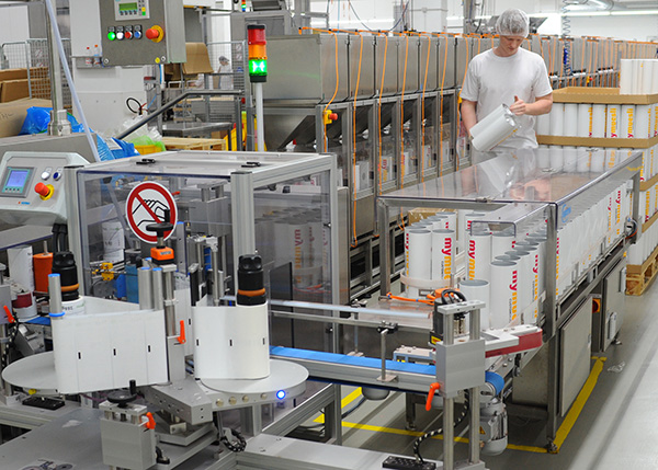 Un travailleur dans une usine de production remplit des conteneurs sur une ligne de remplissage automatisée. Des machines et des emballages sont visibles en arrière-plan.