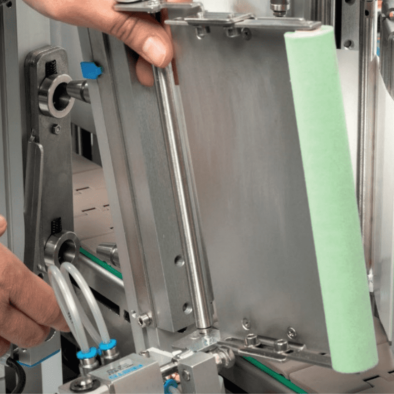 Une main ajuste un dispositif mécanique avec des pièces métalliques et un élément vert. L'environnement montre des équipements industriels.