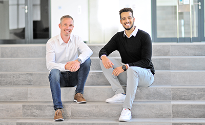 Deux hommes sont assis sur une marche dans un bâtiment moderne. L'un porte une chemise blanche, l'autre un pull noir.