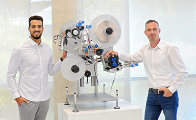 Deux hommes en chemises blanches se tiennent à côté d'une machine technique avec des rouleaux et un écran, conçue pour des applications industrielles.