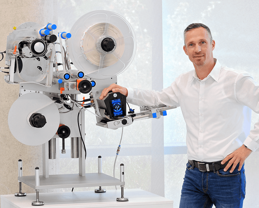 Un homme se tient à côté d'une étiqueteuse moderne et tient un dispositif de commande dans la main. La machine possède plusieurs rouleaux et éléments de contrôle.