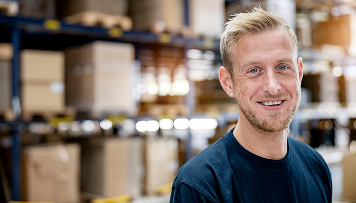 Un homme souriant avec des cheveux blonds se tient dans un entrepôt avec des étagères remplies de cartons en arrière-plan.