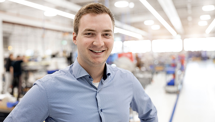 Un jeune homme en chemise bleu clair sourit alors qu'il se tient dans un hall d'usine moderne, entouré de machines et d'espaces de travail.