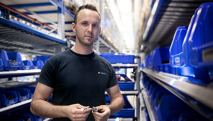 Un homme portant un t-shirt noir tient un outil à la main, entouré de boîtes de rangement bleues sur une étagère de stockage.