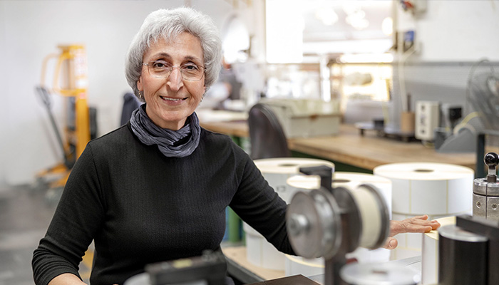 Une femme âgée aux cheveux gris est assise à une table dans un atelier, entourée de rouleaux et de machines. Elle sourit chaleureusement.
