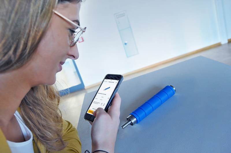 Une personne portant des lunettes regarde son smartphone tout en observant un cylindre bleu sur une table.