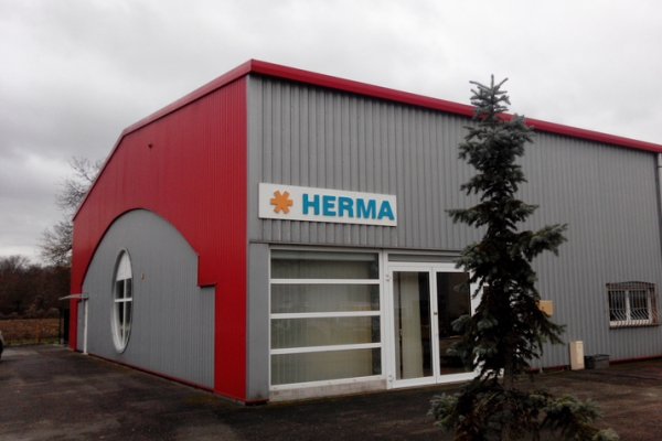 Façade d'un bâtiment moderne avec un toit rouge et des murs gris, portant l'inscription "HERMA". Un arbre se trouve au premier plan.
