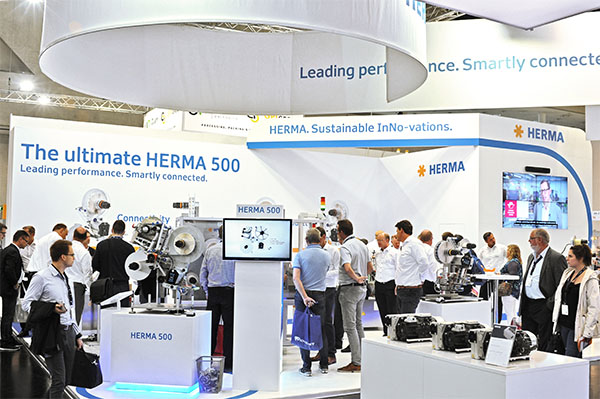 Texte : "Stand de HERMA avec la présentation du HERMA 500. Les visiteurs interagissent avec les machines exposées et les informations.