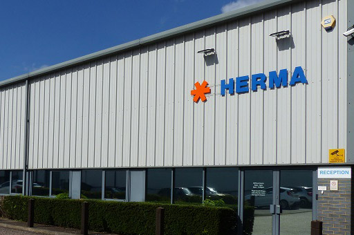 Texte : "Façade d'un bâtiment moderne avec l'inscription "HERMA" en lettres bleues et un symbole orange. Zone d'accueil visible.