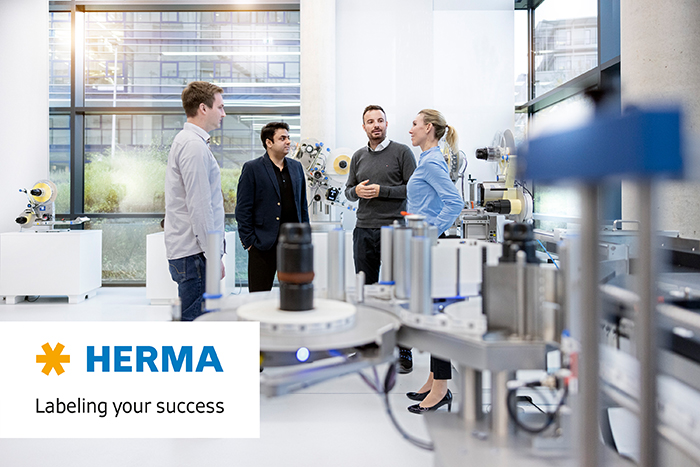 Quatre personnes dans une salle moderne discutent, entourées de machines et d'appareils d'étiquetage. Un logo de HERMA est visible.