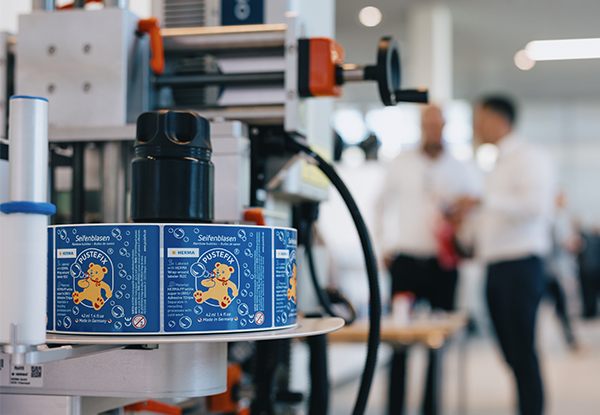 Une machine d'emballage avec des étiquettes colorées sur des canettes au premier plan, tandis que des personnes discutent en arrière-plan.