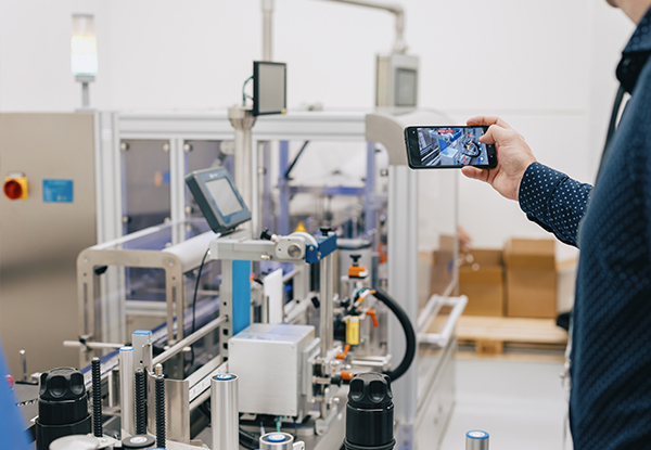 Une personne tient un smartphone et filme une installation de machines automatisées dans un environnement de production.