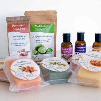 Étiquettes sur différents produits cosmétiques : Doypacks avec du lait de bain et de la poudre de bain, bouteilles en verre brun avec des arômes de bain, ainsi que des savons à l'huile végétale avec des étiquettes rondes dans des sachets transparents, sur fond clair.
