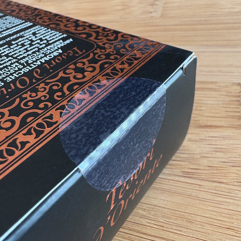 Solutions Cosmétiques : Emballage noir avec des motifs et du texte décoratifs en orange. La languette de l'emballage est scellée avec une étiquette de fermeture transparente.