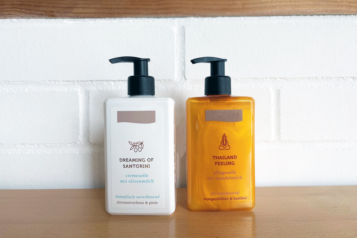 Deux flacons avec pompe contenant du savon crème : un flacon blanc avec une étiquette blanche et un flacon orange avec une étiquette en film transparent au look sans étiquette, posés sur une surface en bois devant un mur en briques blanches.