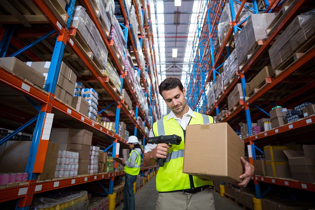 Étiquettes d'expédition et de palettes : Un employé d'entrepôt scanne un carton de transport, avec des rayonnages en hauteur en arrière-plan.