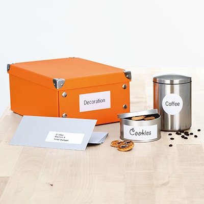 Étiquettes universelles imprimées sur une boîte orange, un récipient à café argenté, une boîte de cookies et une enveloppe grise sur une table en bois.