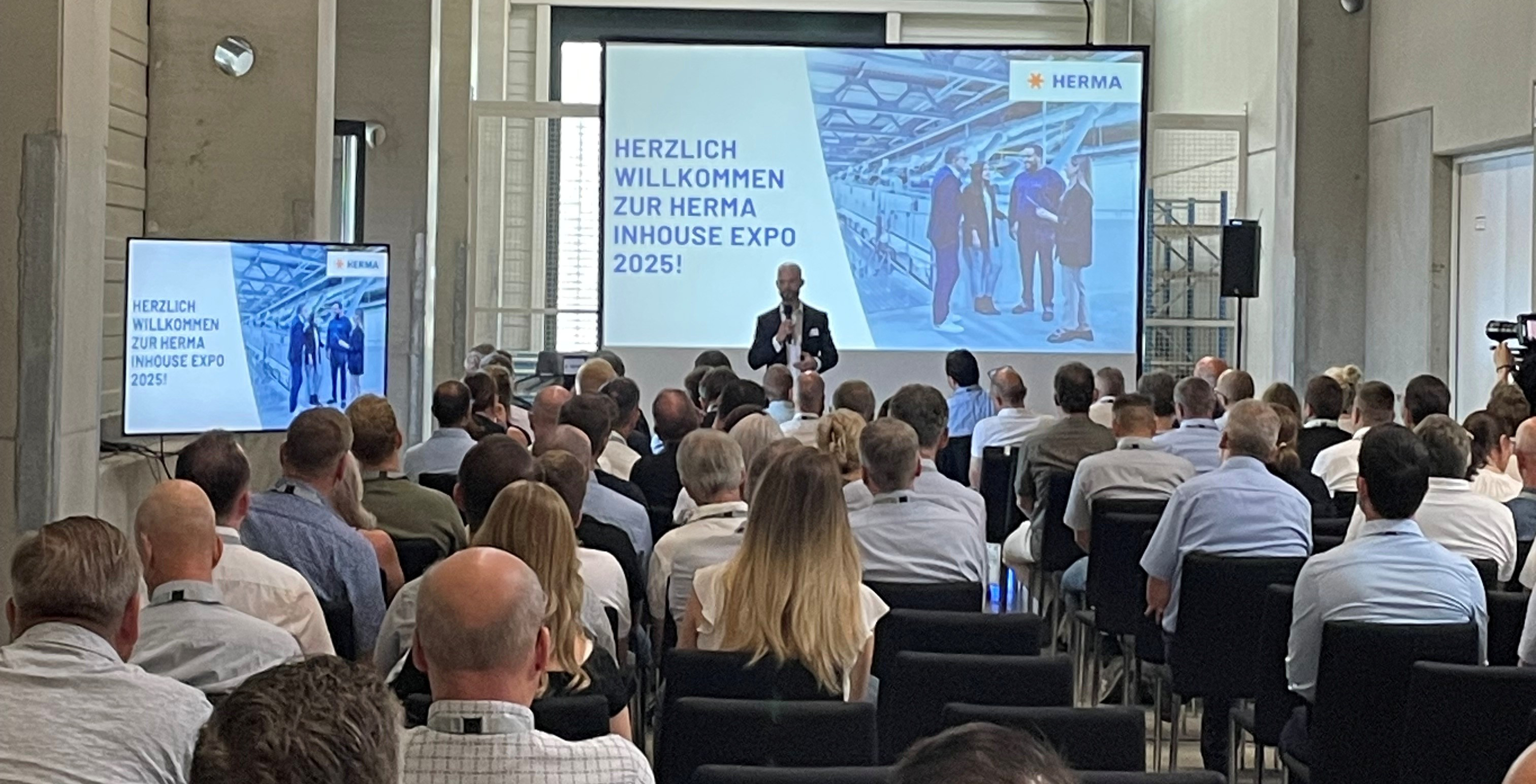 Un orateur présente lors de l'Expo Inhouse HERMA 2025 devant un public. En arrière-plan, on peut voir une diapositive avec le message de bienvenue.