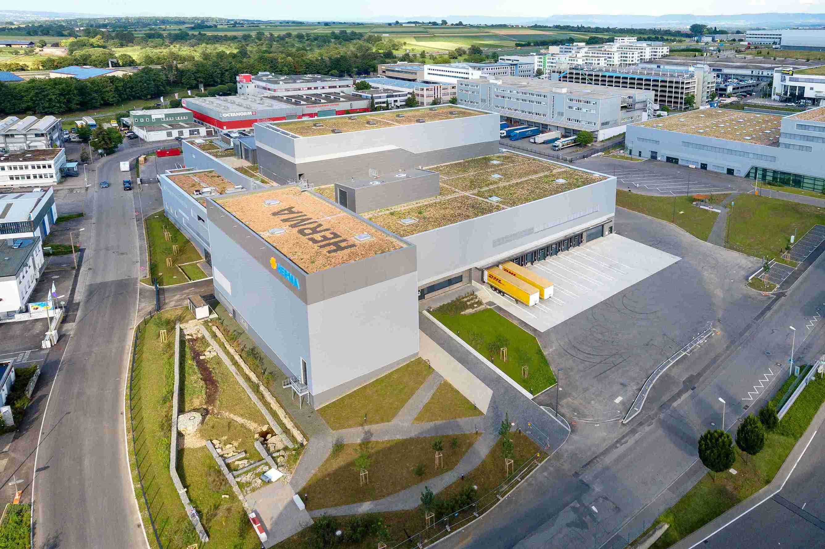 Vue aérienne d'un bâtiment industriel moderne avec un toit végétalisé et des parkings environnants, faisant partie d'une zone d'activité plus vaste.