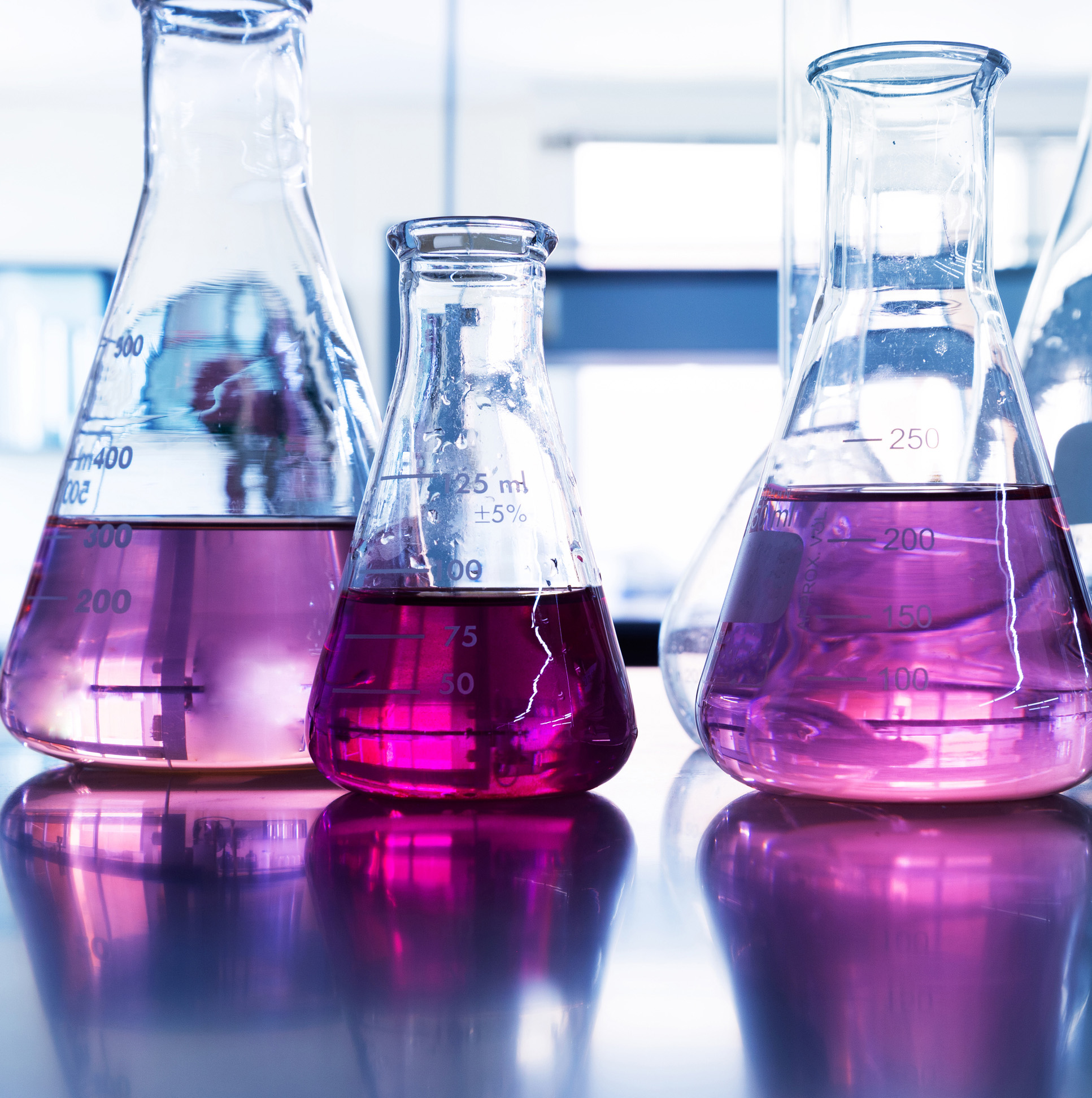 Quatre flacons d'Erlenmeyer contenant un liquide rose, étiquetés avec des échelles de mesure, sur une table dans le laboratoire.