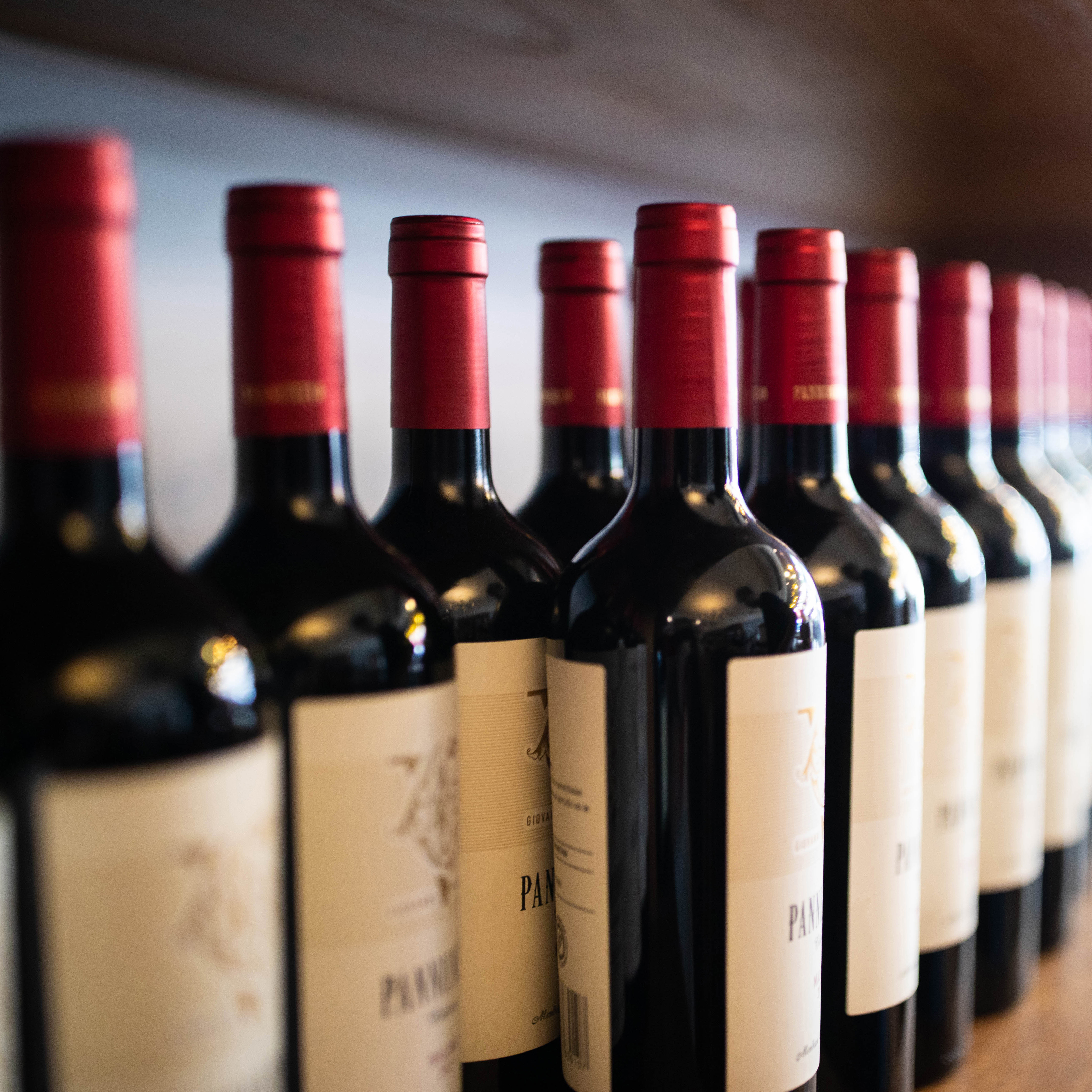 Une rangée de bouteilles de vin rouge avec des capsules rouges, étiquetées et disposées sur une étagère.