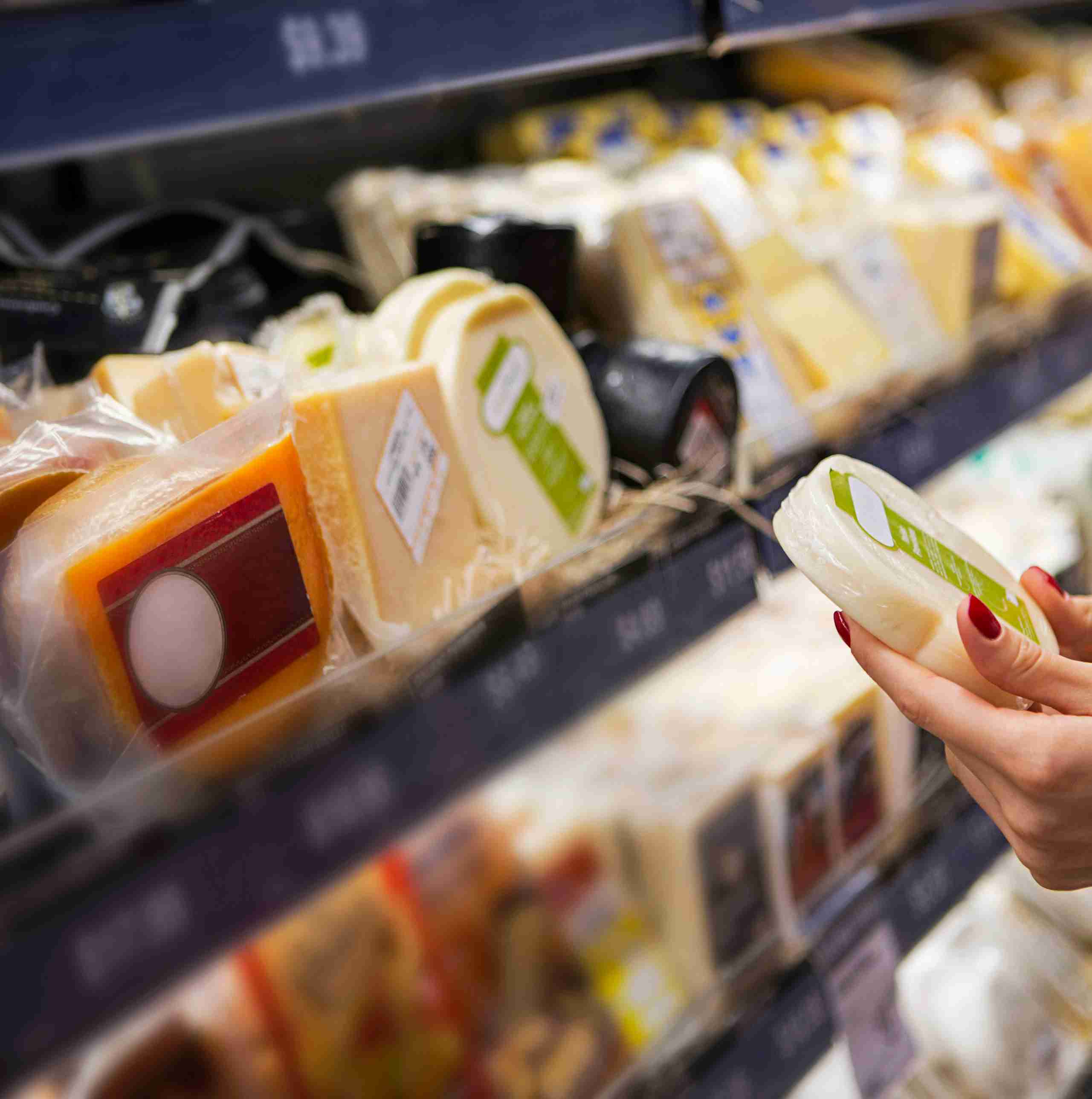 Une main tient un morceau de fromage devant une étagère avec différentes variétés de fromage présentées dans des emballages.