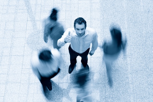 Un homme en chemise blanche fait un signe de pouce levé, entouré de personnes floues qui se déplacent.